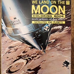 Könnte beinhalten: Ein Vintage-Malbuchcover mit einem Raumschiff, das auf dem Mond landet. Das Cover zeigt eine detaillierte Illustration des Raumschiffs und der Mondoberfläche. Der Titel des Buches lautet "We Land on the Moon Coloring Book" und basiert auf dem NASA-Projekt Apollo. Das Buch ist ein Happiness-Malbuch.