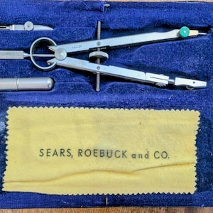 Puede incluir: Un estuche de terciopelo azul abierto que contiene un compás de dibujo plateado, un pequeño cilindro plateado y otras dos herramientas plateadas. Un paño amarillo con el texto "SEARS, ROEBUCK and CO." está en la parte inferior del estuche.