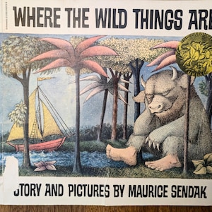 May include: A well-loved copy of "Where the Wild Things Are" by Maurice Sendak. The cover shows a fantastical illustration of a horned, furry creature in a forest setting, with a sailboat and palm trees. The title is in bold, black lettering.