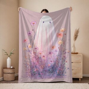 May include: A pink blanket featuring a whimsical ghost design. The ghost is depicted with a white sheet and purple eyes, surrounded by colorful flowers in shades of pink, purple, and yellow. The blanket has a soft, watercolor-like aesthetic.