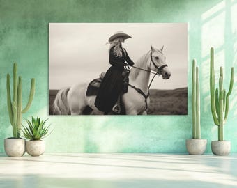 May include: A black and white photograph of a woman wearing a cowboy hat riding a white horse. The woman is wearing a long black dress and the horse is standing in a field.
