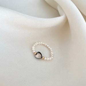 Puede incluir: Un delicado anillo de perlas con un dije en forma de corazón. El dije está hecho de un material gris oscuro con un acento dorado.