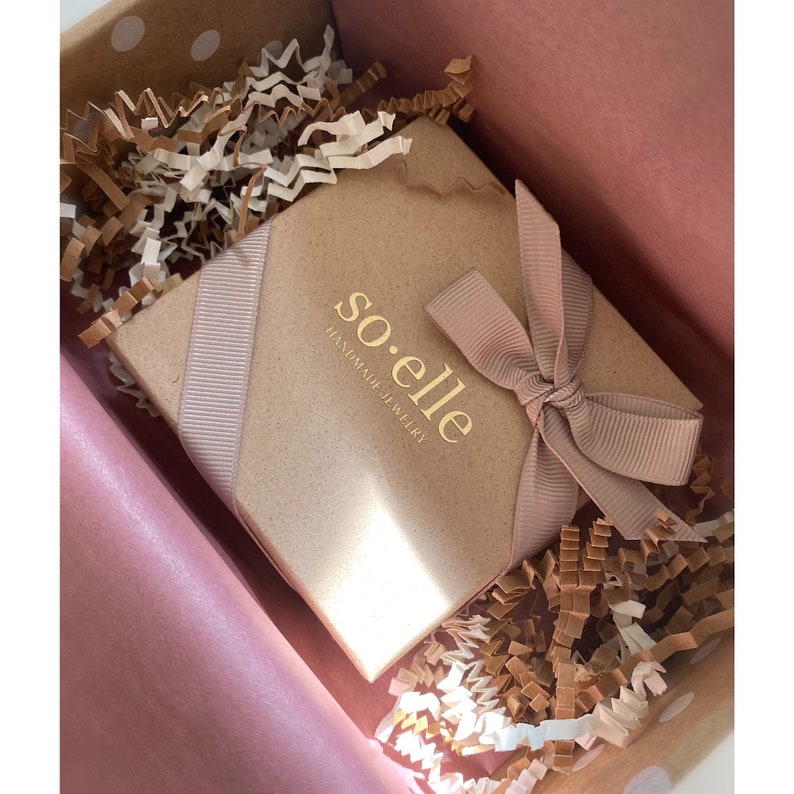 Puede incluir: Una caja de regalo marr&oacute;n con una cinta rosa y el texto "so.elle handmade jewelry" en letras doradas. La caja est&aacute; rodeada de papel arrugado marr&oacute;n y blanco.
