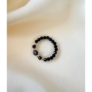 以下が含まれることがあります： 黒いファセットビーズとゴールドカラーのアクセントが特徴のビーズリング。リングには、青と白の邪視のデザインが施された中央のチャームが含まれており、ジュエリーにユニークで象徴的な要素を追加しています。