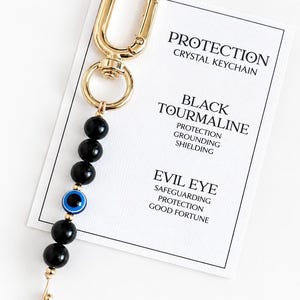 May include: A gold-toned metal keychain featuring five round black beads and one blue, white, and black evil eye bead. The keychain is attached to a white display card printed with the text PROTECTION CRYSTAL KEYCHAIN, BLACK TOURMALINE, and EVIL EYE.