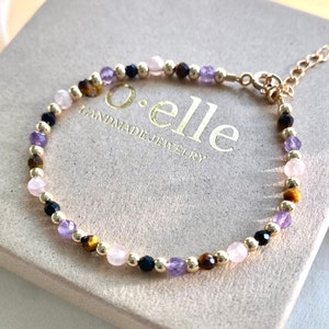 Könnte beinhalten: Ein Perlenarmband mit abwechselnden facettierten Edelsteinen in Violett, Rosa, Schwarz und Braun, durchsetzt mit kleinen goldfarbenen Metallkugeln. Das Armband verfügt über eine goldfarbene Metall-Verlängerungskette und einen runden Verschluss.