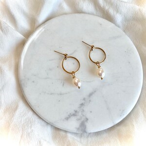 May include: A pair of gold hoop earrings with a single white pearl dangling from each hoop.