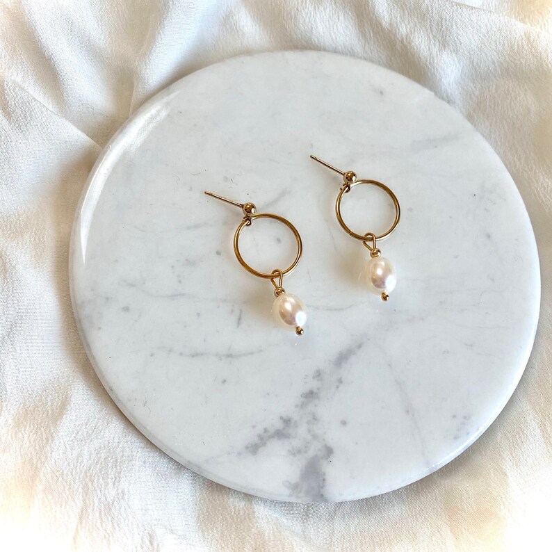 May include: A pair of gold hoop earrings with a single white pearl dangling from each hoop.