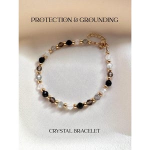 May include: A beaded bracelet featuring alternating black, clear, and smoky gray faceted crystal beads interspersed with small gold-toned metal spheres. The bracelet includes a gold-toned metal chain extender and clasp. Text reads PROTECTION & GROUNDING CRYSTAL BRACELET.