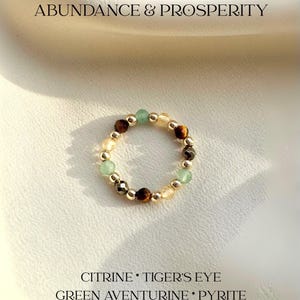 May include: A beaded ring featuring alternating citrine, tiger's eye, green aventurine, and pyrite gemstones with small gold-tone metallic beads. The text ABUNDANCE & PROSPERITY and CITRINE * TIGER'S EYE GREEN AVENTURINE * PYRITE is visible.