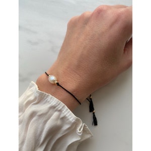 May include: A black string bracelet with a single white pearl and gold accents. The bracelet has black tassels on the ends.