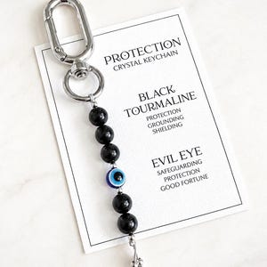 May include: A keychain featuring black tourmaline beads and a single blue, white, and black evil eye bead attached to a silver-toned metal clasp. The keychain rests on a white card printed with the text PROTECTION CRYSTAL KEYCHAIN, BLACK TOURMALINE, PROTECTION, GROUNDING, SHIELDING, EVIL EYE, SAFEGUARDING, PROTECTION, GOOD FORTUNE.
