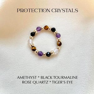 May include: A beaded ring featuring faceted amethyst, black tourmaline, rose quartz, and tiger's eye gemstones interspersed with small gold-toned metallic beads. The text PROTECTION CRYSTALS and AMETHYST * BLACK TOURMALINE ROSE QUARTZ * TIGER'S EYE is visible.