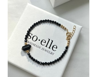 Peut inclure: Un bracelet composé de petites perles noires à facettes et d'une pointe en pierre noire centrale, agrémenté de perles en métal doré. Le bijou comprend un fermoir mousqueton doré et une chaînette réglable, posé sur une boîte blanche marquée so·elle.