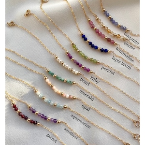 Pode incluir: Uma coleção de delicados colares de ouro, cada um com uma fileira de pedras preciosas coloridas: granada, ametista, água-marinha, opala, esmeralda, pérola, rubi, peridoto, lápis-lazúli, turmalina, citrino e tanzanita. Os nomes das pedras estão rotulados.