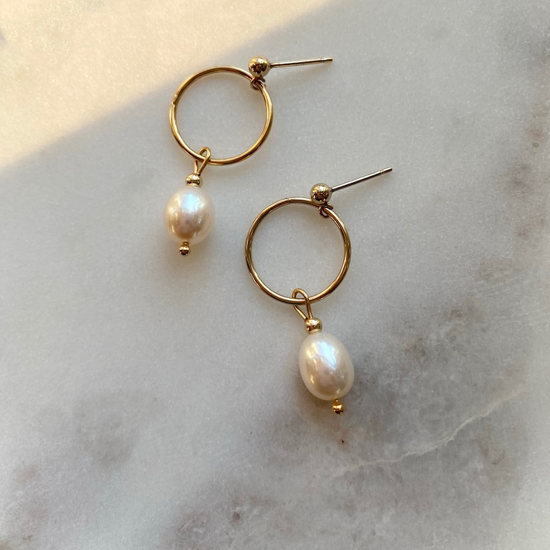 May include: A pair of gold hoop earrings with a single white pearl dangling from each hoop.