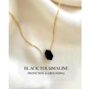 Puede incluir: Un collar de cadena dorada con un colgante negro hexagonal. El collar se muestra sobre un fondo blanco texturizado. El texto "BLACK TOURMALINE PROTECTION & GROUNDING" está en la parte inferior de la imagen.