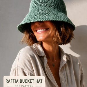 Puede incluir: Un sombrero de pescador tejido a ganchillo en color verde salvia con un ala ancha inclinada hacia abajo y una construcción tejida texturizada. Una etiqueta rectangular en la esquina inferior muestra el texto "RAFFIA BUCKET HAT" y "PDF PATTERN".