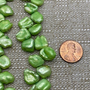 May include: A strand of green freshwater pearls, each pearl is a unique shape and size. The pearls are displayed next to a US penny for size reference.