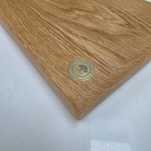 May include: A rectangular wooden cutting board with a natural wood grain finish. A circular brass emblem with the text "Foundry & Forest Est. 2026" is affixed to the board. The board is likely used for food preparation.