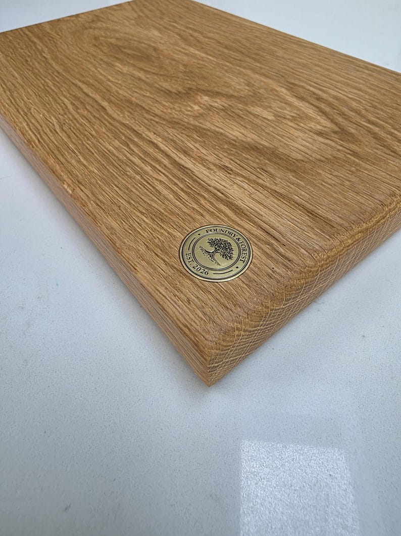 May include: A rectangular wooden cutting board with a natural wood grain finish. A circular brass emblem with the text "Foundry & Forest Est. 2026" is affixed to the board. The board is likely used for food preparation.