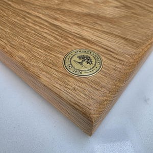 May include: Close-up of a rectangular wooden cutting board with a rounded edge. The board has a natural wood grain pattern and a gold-colored circular emblem with the text "Foundry & Forest Est. 2026".