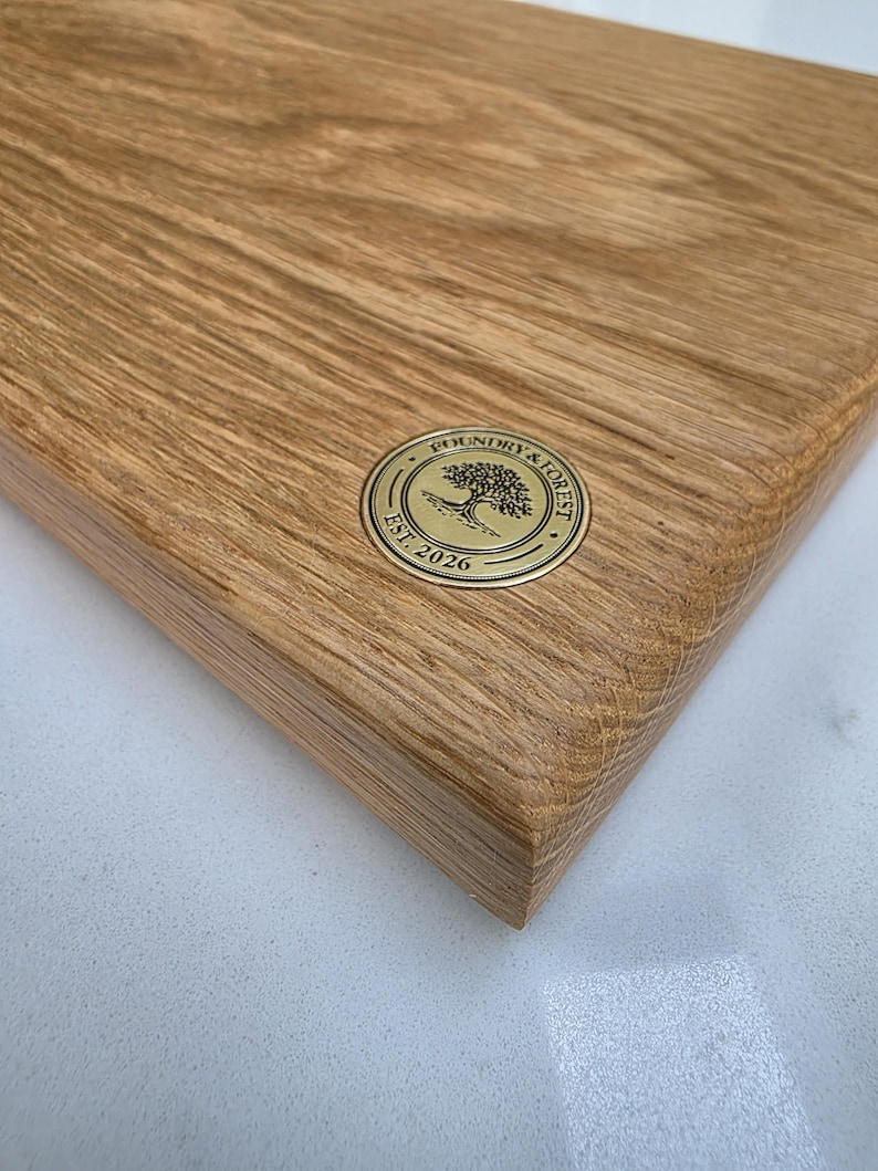 May include: Close-up of a rectangular wooden cutting board with a rounded edge. The board has a natural wood grain pattern and a gold-colored circular emblem with the text "Foundry & Forest Est. 2026".