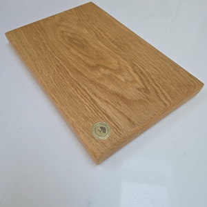 May include: A rectangular wooden cutting board with a light brown finish. The board has a natural wood grain pattern and a small, round, gold-colored emblem in the corner. The board is approximately 12 inches long and 8 inches wide.
