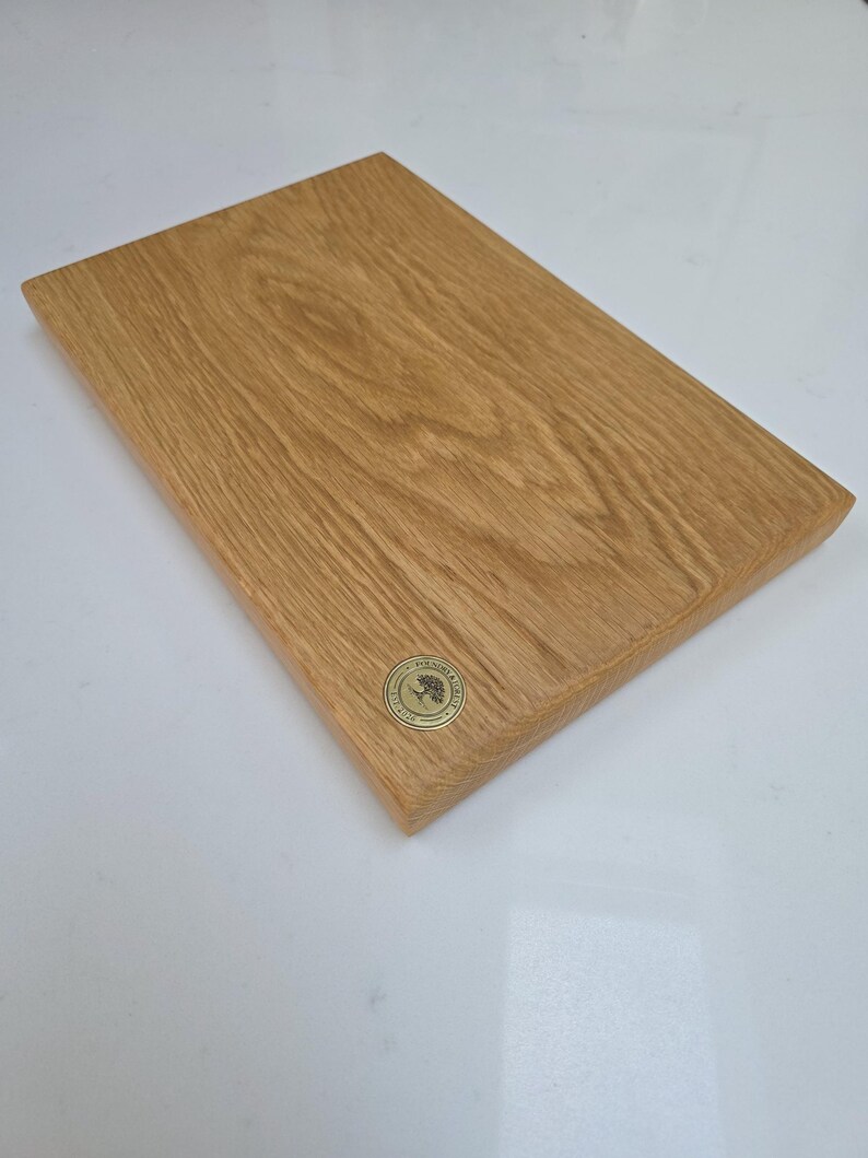 May include: A rectangular wooden cutting board with a light brown finish. The board has a natural wood grain pattern and a small, round, gold-colored emblem in the corner. The board is approximately 12 inches long and 8 inches wide.