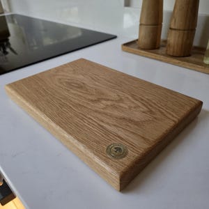 May include: A rectangular wooden cutting board with a light brown finish. The board has a rounded edge and a circular metal emblem in the corner. The board is sitting on a white countertop.