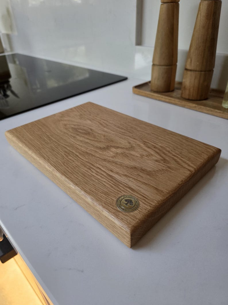 May include: A rectangular wooden cutting board with a light brown finish. The board has a rounded edge and a circular metal emblem in the corner. The board is sitting on a white countertop.