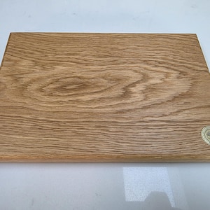 May include: A rectangular wooden cutting board with a light brown finish. The wood grain is visible, and a small, round emblem with a tree design is in the lower right corner. This kitchen item is perfect for food preparation.