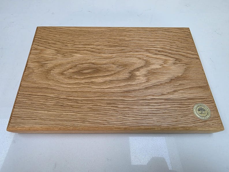 May include: A rectangular wooden cutting board with a light brown finish. The wood grain is visible, and a small, round emblem with a tree design is in the lower right corner. This kitchen item is perfect for food preparation.
