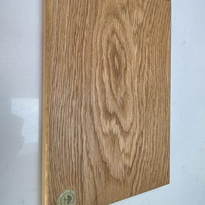 May include: A rectangular wooden cutting board with a natural wood grain pattern. The board is light brown with visible wood grain and a small, circular logo in the lower left corner. The board is displayed against a white background.