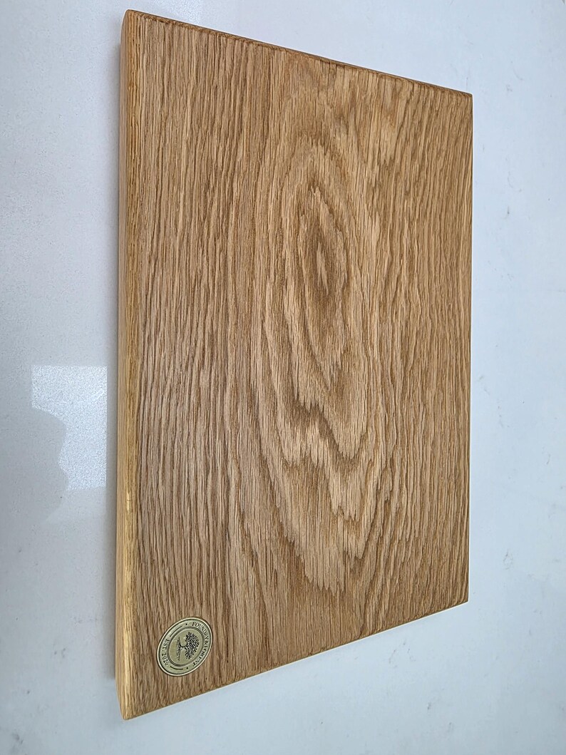 May include: A rectangular wooden cutting board with a natural wood grain pattern. The board is light brown with visible wood grain and a small, circular logo in the lower left corner. The board is displayed against a white background.