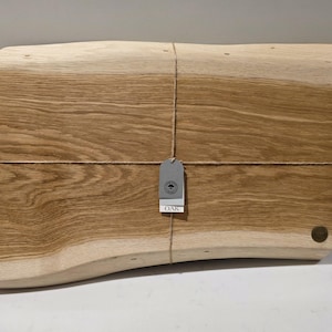 May include: A rectangular wooden cutting board with a natural wood grain pattern. The board is tied with twine and has a small tag with the word "OAK" visible. The board has a curved edge and a small, round, dark-colored accent.
