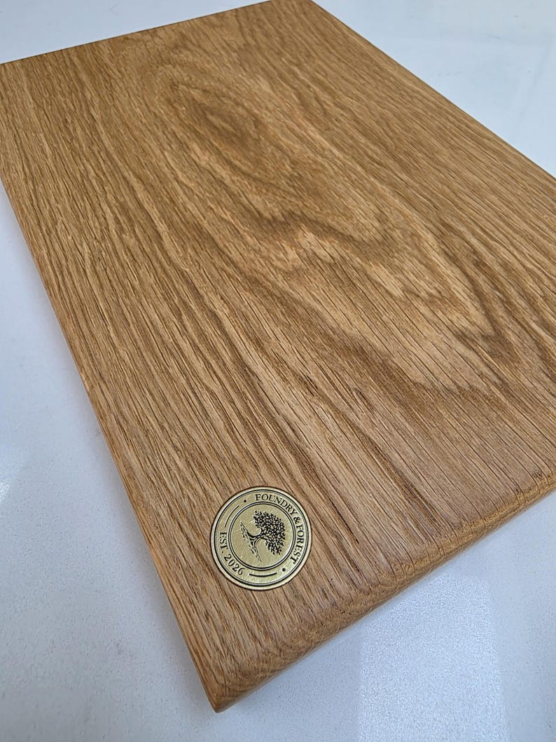 May include: A rectangular wooden cutting board with a natural wood grain finish. A circular brass emblem with the text "Foundry & Forest Est. 2026" is affixed to the board. The board is likely used for food preparation.