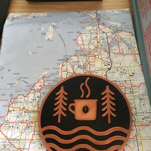 Op de afbeelding: Een ronde, koperkleurige onderzetter met een zwarte achtergrond met een koffiekopje, bomen en golvende lijnen. De onderzetter ligt op een kaart van het Traverse Bay-gebied.