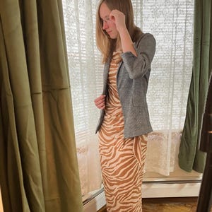 Puede incluir: Un blazer gris sobre un vestido largo con estampado de cebra marrón y beige. El vestido tiene una silueta ajustada. El blazer tiene un corte clásico con mangas tres cuartos. El fondo incluye una ventana con cortinas de encaje y cortinas verdes.