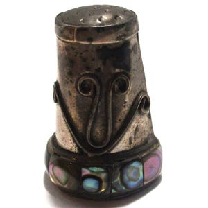 May include: A vintage silver thimble with a decorative face design. The thimble features a series of holes on the top and is adorned with iridescent abalone shell accents around the base. The thimble is set against a white background.