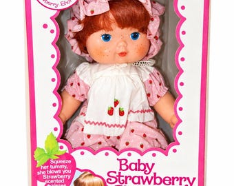May include: A Baby Strawberry Shortcake doll in a pink box. The doll has red hair, blue eyes, and freckles, wearing a pink strawberry-print dress and bonnet. The box features the text Welcome to the World of Strawberry Shortcake and Baby Strawberry Shortcake.