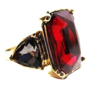 May include: A gold-toned ring featuring a large, rectangular, deep red gemstone and a smaller, triangular, dark gemstone. The ring has an ornate setting with intricate details, showcasing a bold and elegant design.