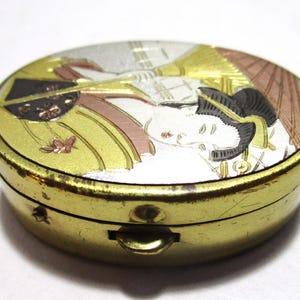 May include: A small, oval-shaped, antique pillbox with a gold-colored exterior. The lid features a detailed illustration of a woman in traditional Japanese attire, with gold, silver, and bronze accents. The box has a small clasp.