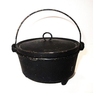 May include: A black cast iron Dutch oven with a lid and a wire handle. The pot has three small feet and a loop handle on the lid. The pot is designed for cooking.