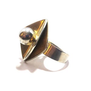 May include: A silver ring featuring a large, diamond-shaped centerpiece with a smooth, amber-colored inlay. A polished silver sphere is mounted on the upper portion of the diamond shape, which is attached to a simple, wide silver band.