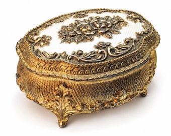 May include: An oval-shaped jewelry box featuring a metallic gold finish with intricate embossed leaf and lattice patterns. The lid displays a white textured center panel adorned with a raised gold floral relief and a decorative beaded border.