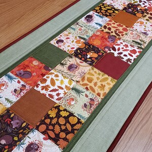 May include: A rectangular table runner featuring a patchwork design of squares with autumn-themed prints including hedgehogs, foxes, and fall leaves in shades of orange, brown, and cream, bordered by solid green and dark red fabric strips.