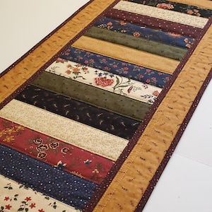 May include: A rectangular fabric table runner featuring a patchwork design of alternating horizontal strips. The strips display various floral and geometric patterns in shades of navy blue, burgundy, olive green, cream, and gold, bordered by a gold trim.