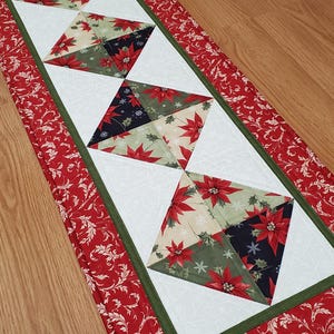 May include: A rectangular quilted table runner featuring a central white panel with a subtle snowflake pattern. Four diamond-shaped blocks are arranged in a row, each composed of fabric squares with red poinsettias, green holly, and dark navy blue.