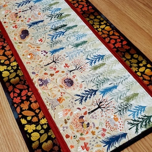 May include: A rectangular table runner featuring a central panel with a cream background, decorated with illustrations of trees, squirrels, hedgehogs, and mushrooms. It is bordered by a red fabric strip and a black fabric edge with autumn leaves.